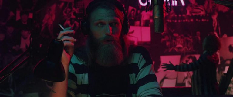 A bearded man, wearing a striped black and white top, sits in front of a radio microphone holding a cigarette. He is bathed in blue and red lighting and in the background are vague horror and band posters.