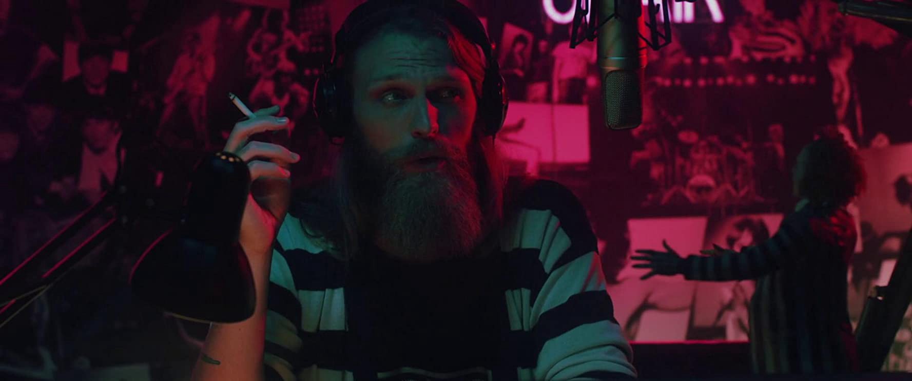 A bearded man, wearing a striped black and white top, sits in front of a radio microphone holding a cigarette. He is bathed in blue and red lighting and in the background are vague horror and band posters.