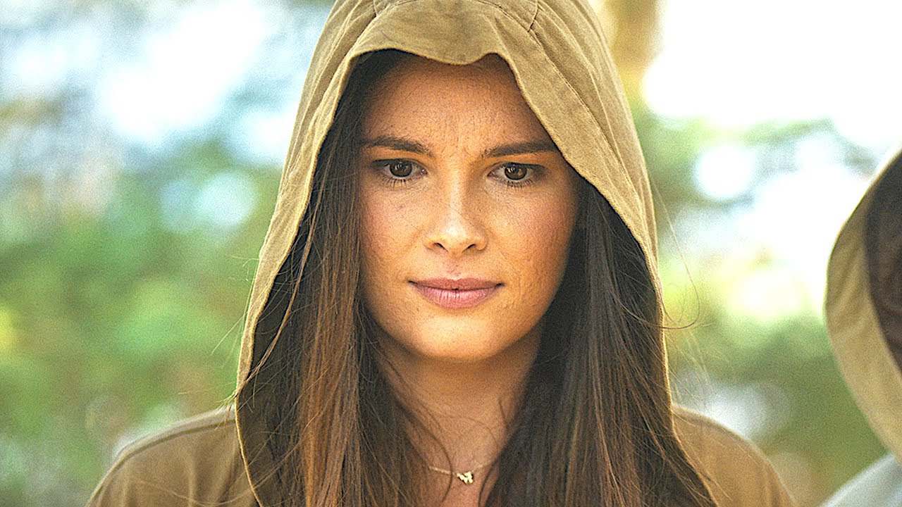 A woman wearing a brown hooded raincoat, looks down with a concerned expression. Behind is a treeline.