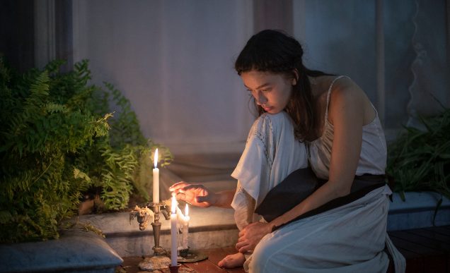 Screenshot from 'Glasshouse'. A woman wearing white garments sits in thought by some candles.