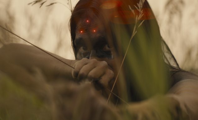 Screenshot from 'Prey'. A Comanche warrior hides in tall grass aiming with a bow, whilst three laser dots aim for his head.