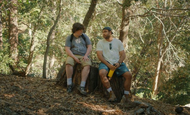 A teenager and a man, wearing hiking gear, sit on a log together in a forest on the mountain