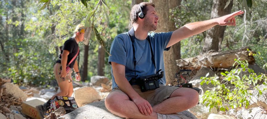 A film director, Corey Sherman, sits cross-legged and points out towards someone in a wooded area.