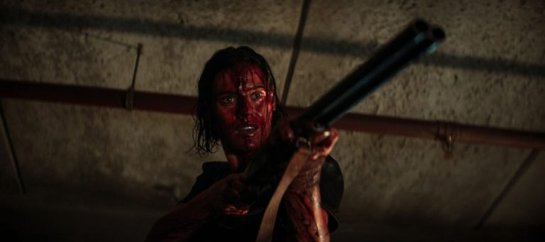 A blood-soaked women aims a shotgun at something off screen
