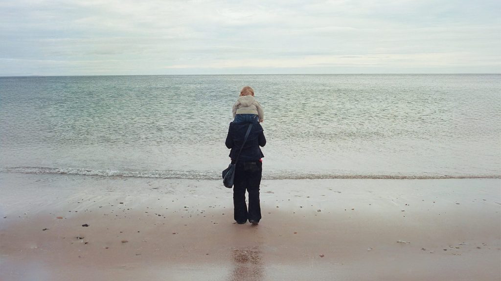 A mother has her young son sat on her shoulders. They're at the beach, looking out towards the sea