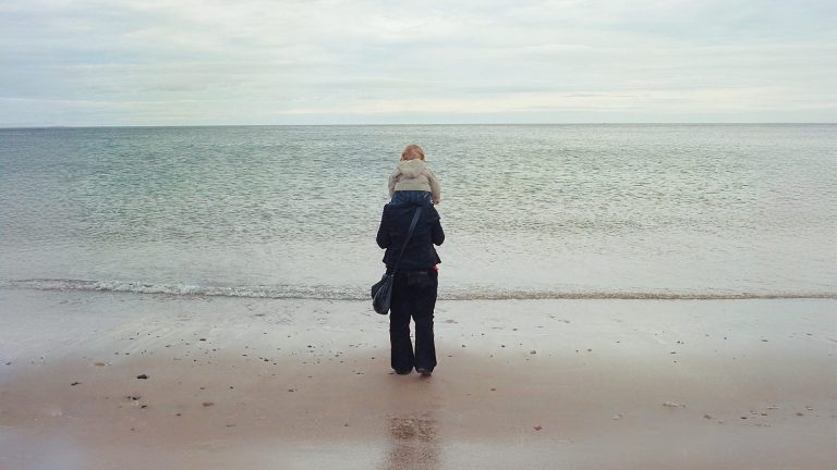 A mother has her young son sat on her shoulders. They're at the beach, looking out towards the sea