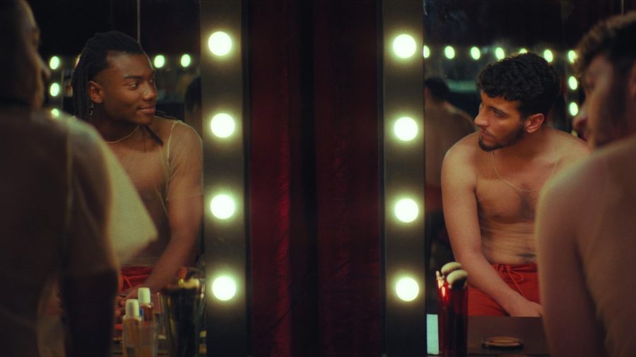 Two topless men sat in front of illuminated mirrors in a scene from Night Stage