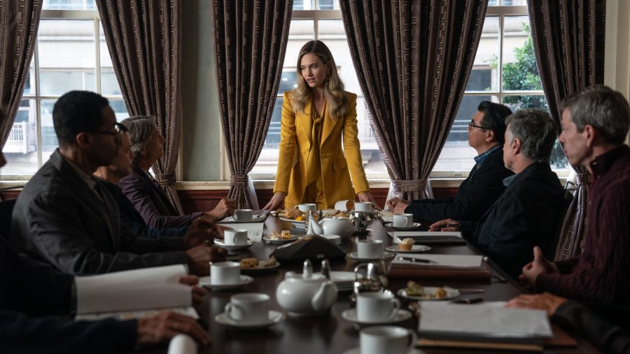 Lily James stands at the head of a table with men sat either side of her,