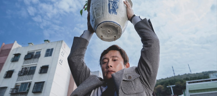 Lee Byung-hun as Yoo Man-soo lifts a heavy flower pot over his head with thoughts of mischief in No Other Choice