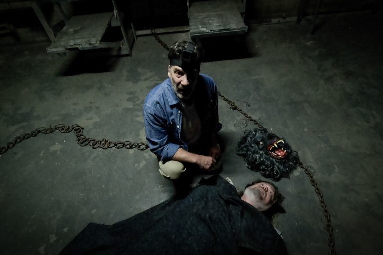 Mark Duplass as Josef standing over the body of a man next to a werewolf mask in a scene from The Creep Tapes.