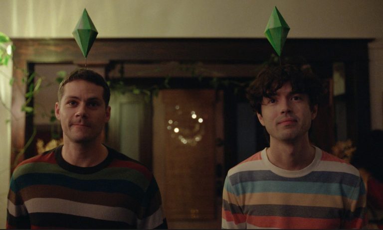 Characters Roman and Dennis stand at the doorway of a home dressed as 'Sims' with green diamonds above their heads. Both are wearing striped jumpers. This is a still from the film.