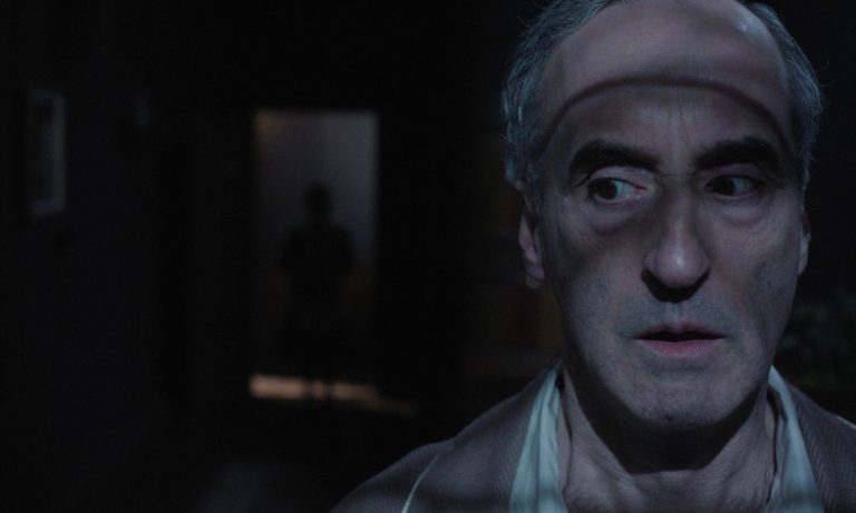 Don McKellar as Minturn standing in a dark room with shadows across his face in a scene from The Occupant of the Room.